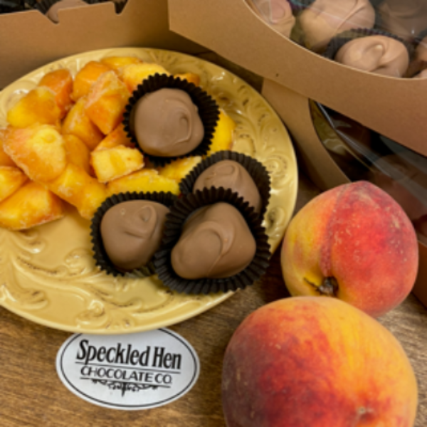 A close-up of assorted chocolate candies and peach halves arranged on a tabletop, with cardboard boxes of more chocolates in the background. A round logo sticker reads 'Spckled Hen Chocolate Co'.